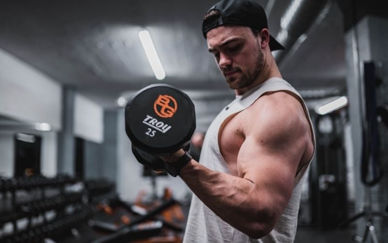 Muscular man in white tank top doing bicep curls with a 25 pound dumbbell.
