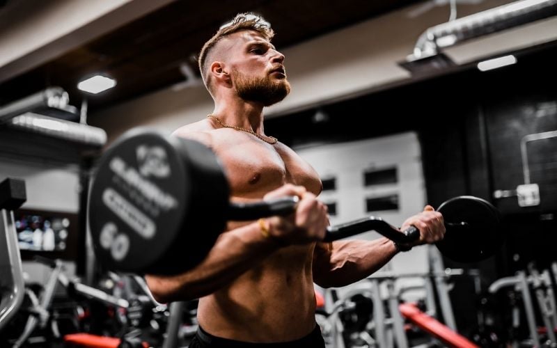 Muscular, shirtless man doing EZ bar bicep curls with 60 pound dumbbell.