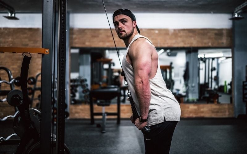 Muscular man in white tank and black backwards hat doing rope tricep pushdowns.
