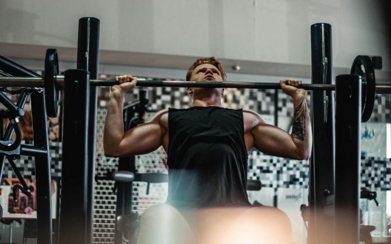 Muscular man in black tank top prepping for seated barbell shoulder press.