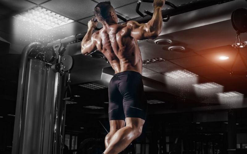 Shirtless man in black shorts doing pullups in gym.