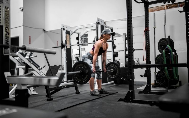 Woman in leggings, sports bra, and hat dong deadlifts in a gym.