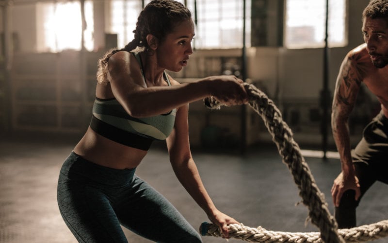 Woman doing battle ropes in gym setting with man cheering her on.