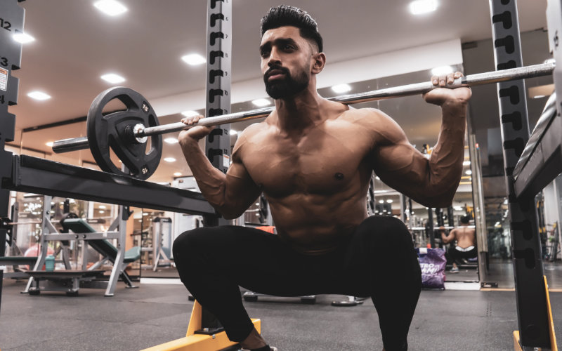 Shirtless man doing barbell back squats in gym.