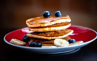Stack of pancakes topped with blueberries and bananas and drizzled with syrup on a red plate.