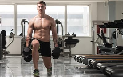 Muscular man holding dumbbells while doing dumbbell walking lunges.