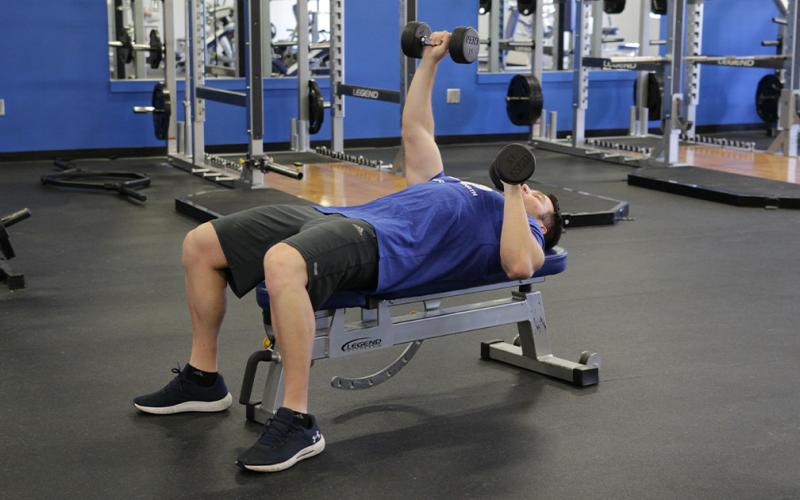 Fit man doing an alternating dumbbell bench press in the gym.