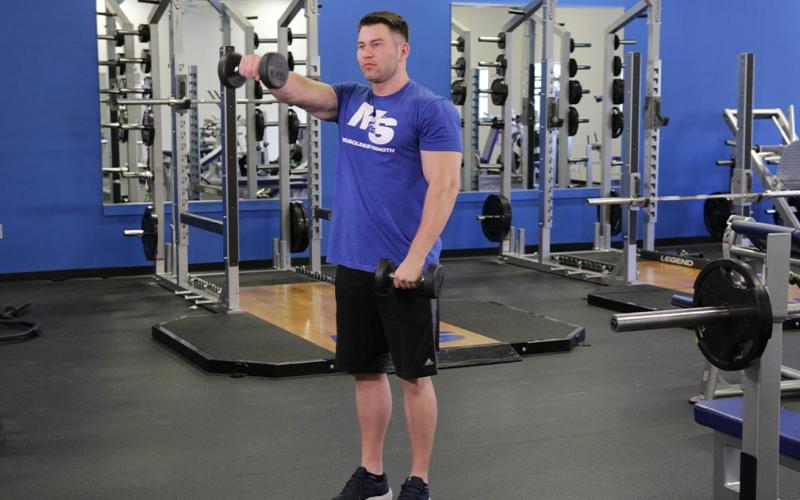 Fit man doing an alternating dumbbell front raise in the gym.