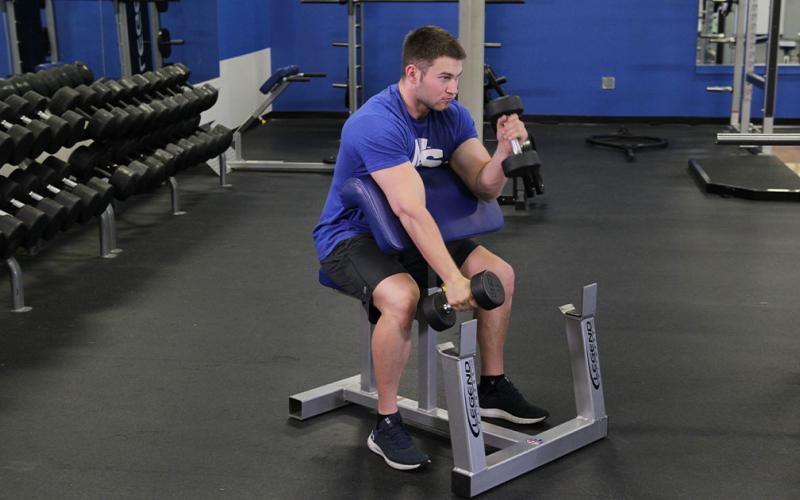 Fit man doing an alternating dumbbell hammer preacher curl in the gym.
