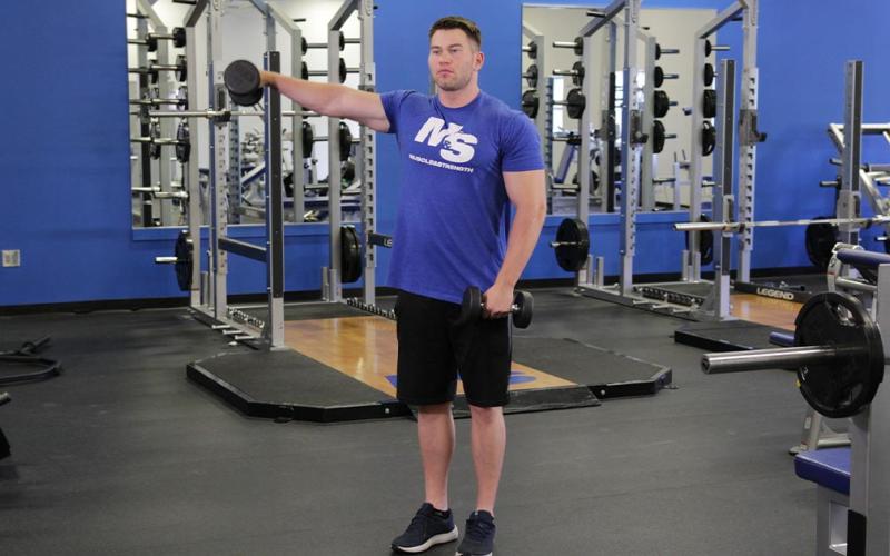 Fit man doing an alternating dumbbell lateral raise in the gym.
