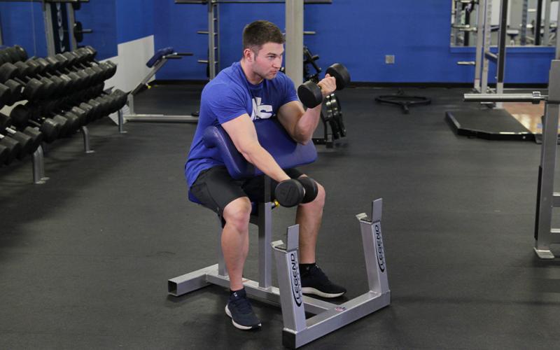 Fit man doing an alternating dumbbell preacher curl in the gym.