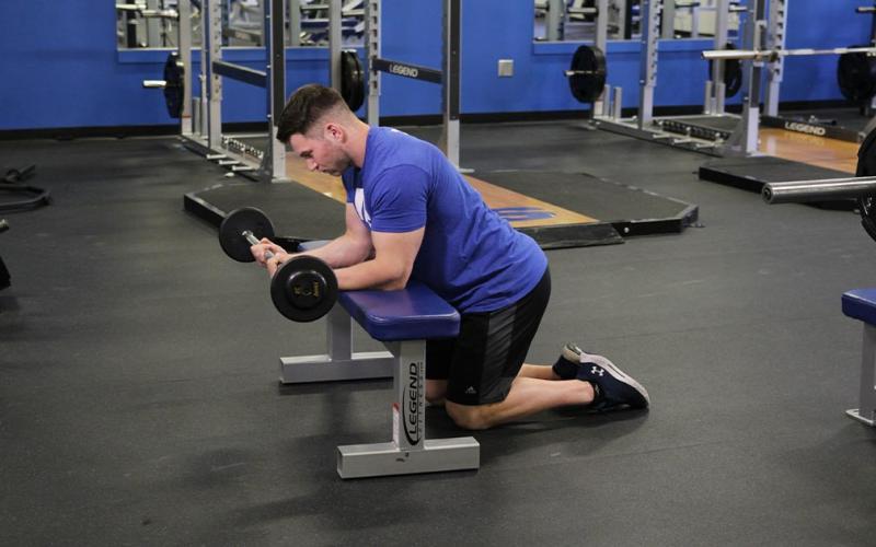 Fit man doing a barbell wrist curl over a bench in the gym.