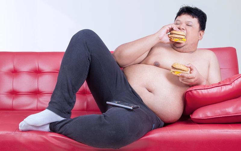 Close up shot of a large shirtless man eating two burgers on the couch.