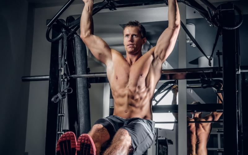 Shirtless muscular man doing abs workouts on pull up bar in a gym.