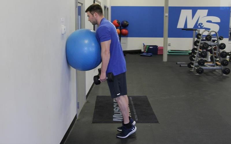 Fit man doing a calf raise while leaning against an exercise ball on the wall.
