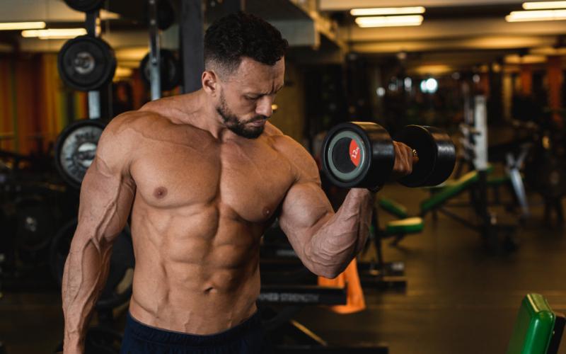 Muscular man doing alternating dumbbell bicep curl in gym.