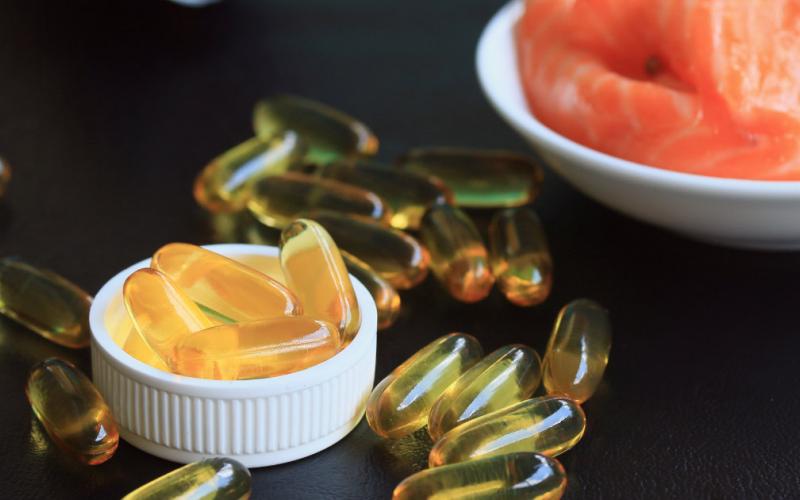 Yellow fish oil capsules on a black table in front of a bowl of raw salmon.