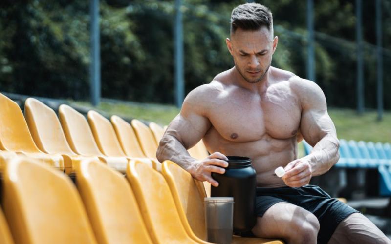 Shirtless muscular man making a post-workout protein shake outside.