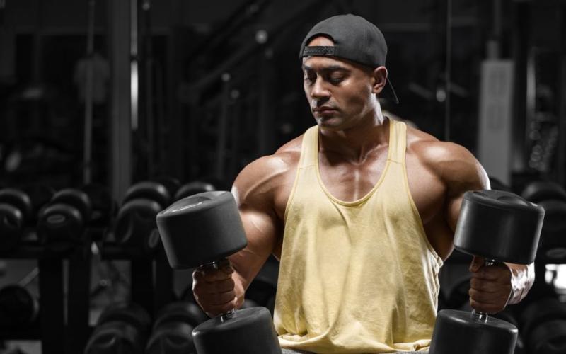Muscular man wearing a yellow tank top holding dumbbells in the gym.