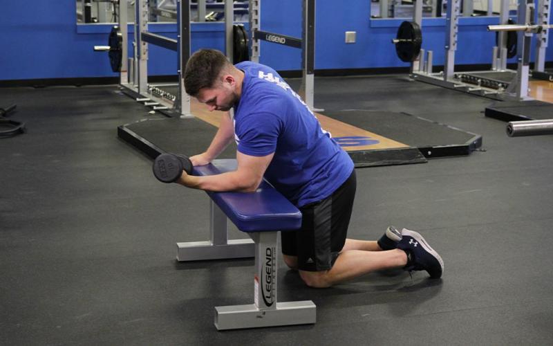 Fit man doing a one-arm dumbbell wrist curl over bench in the gym.