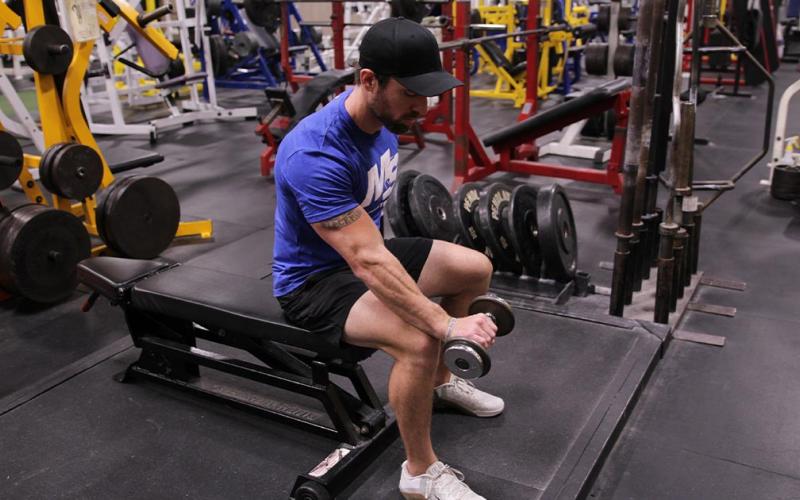 Fit man doing a one-arm seated reverse grip dumbbell wrist curl in the gym.