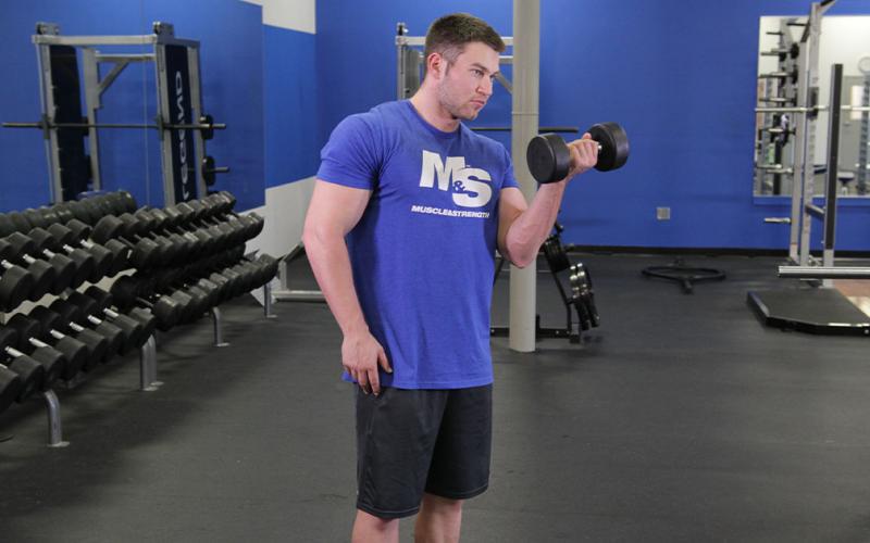 Fit man doing a one-arm standing dumbbell curl in the gym.