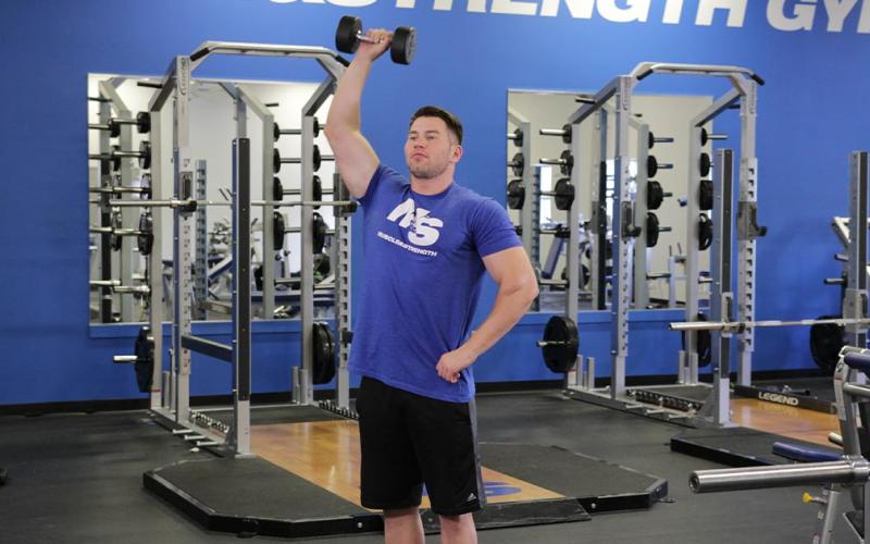 Fit man doing a one-arm standing dumbbell shoulder press in the gym.