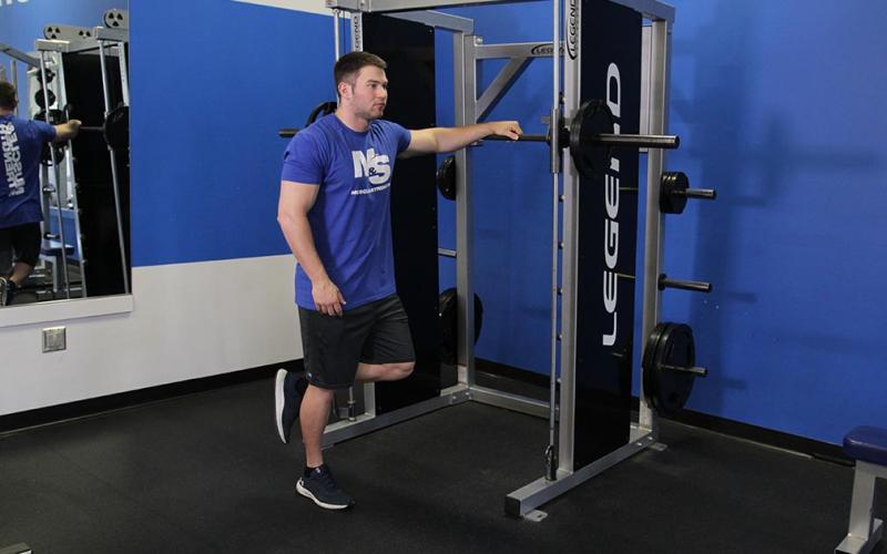 Fit man doing a one-leg calf raise on the floor while holding onto a smith machine.