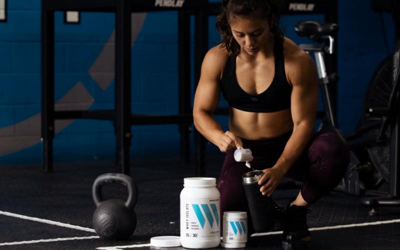 Swolverine athlete scooping whey protein into shaker on floor of CrossFit-style gym.