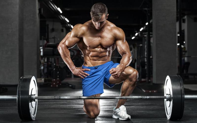 Shirtless, muscular man posing on one knee in front of barbell.