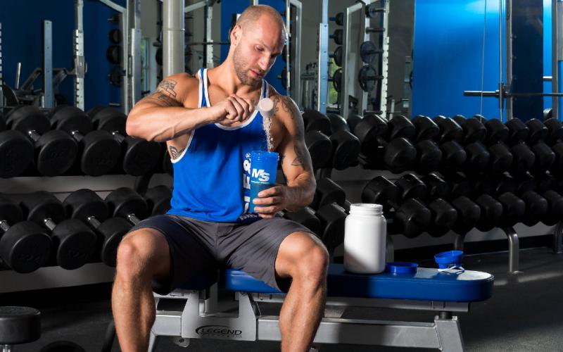 M&S model sitting on bench in gym while scooping protein powder into shaker cup.