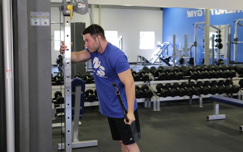 Fit man doing a reverse one-arm cable tricep extension at the gym.