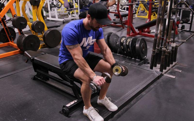 Fit man doing a Seated Reverse Grip Dumbbell Wrist Curl in the gym.