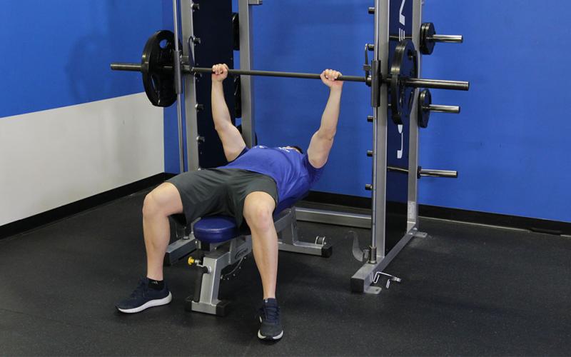 Fit man doing a smith machine wide grip bench press in the gym.