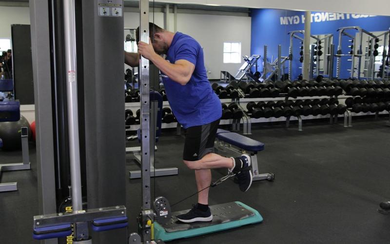 Fit man doing a standing cable hamstring curl in the gym.