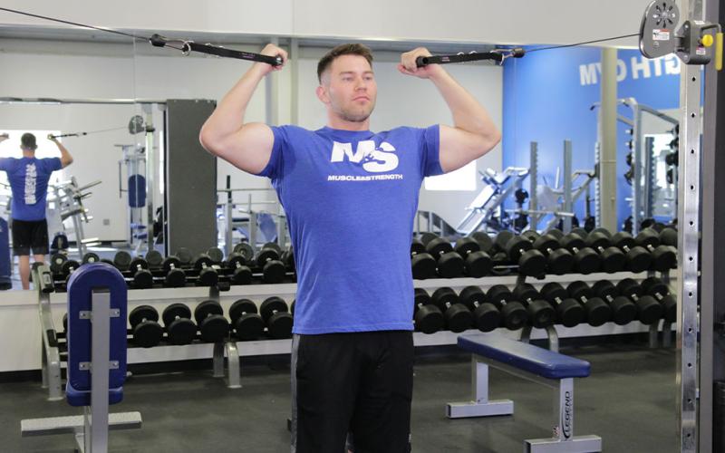 Fit man doing a standing high pulley cable curl in the gym.