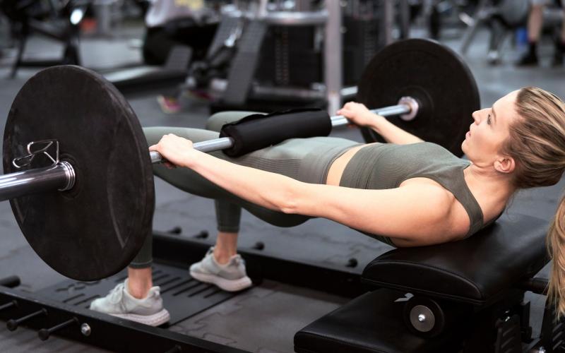 Strong blonde woman doing barbell hip thrusts in the gym.