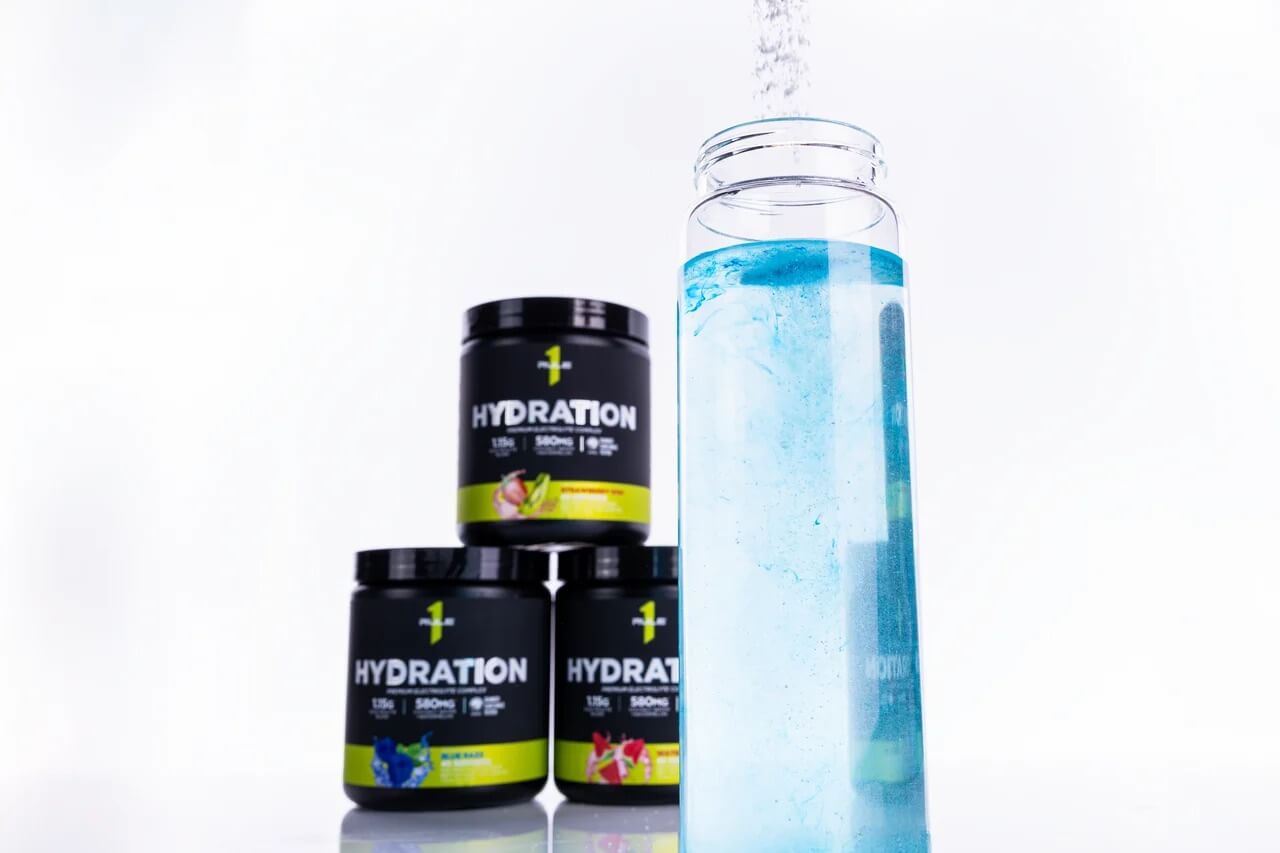 Three containers of Rule 1 Hydration powder in various flavors stacked with a clear glass filled with blue electrolyte solution